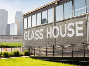 Photo of the Glass House. The front wall is featured with gray brick and white lettering that reads: "GLASS HOUSE". Green grass lines the front of the building up to a long ramp that runs along the wall. In the background is part of the Minneapolis skyline.