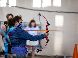 Shinoa, wearing a blue coat and a mask, taking part in an archery activity with Lifeworks.