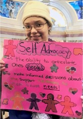 Shinoa at the state capitol building in Saint Paul with a pink sign that reads: "Self Advocacy is the ability to articulate ones needs. To make informed decisions about the support necessary to meet those needs!"