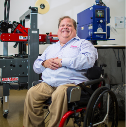 A photo of a man wearing a light blue shirt and khaki pants. He is sitting in a wheelchair in front of various industrial equipment.