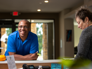 A photo of a man in a blue polo shirt leaning on a desk and smiling.