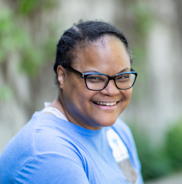 A photo of a woman wearing a blue t-shirt and glasses. She is smiling and sitting in front of an outdoor wall covered in green foliage.